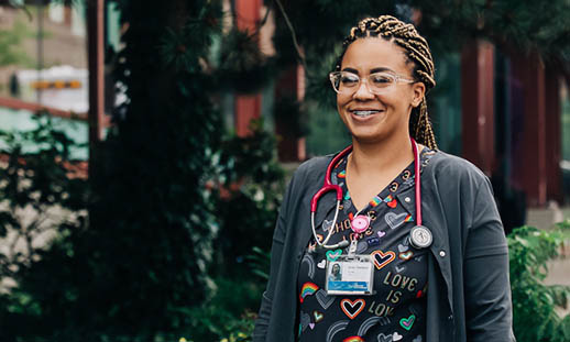 A woman, wearing scrubs, a badge and a stethoscope around her neck, walks in front of the QE2 Hospital. She is smiling broadly.