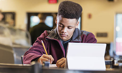 A man sits at a table and writes with a pencil. He is wearing a maroon sweater and has his laptop open in front of him.