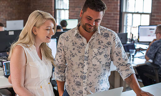 A blond woman and a bearded man stand shoulder to shoulder at a desk and look at an Apple laptop. There is a brick-walled office space in the background with several desks and people.