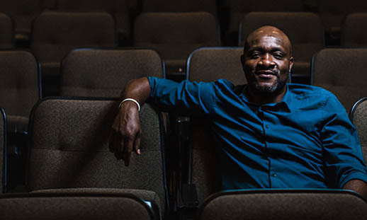 A man sits in an empty row of an auditorium. He is wearing a blue, button up shirt and looks at the camera with a smirk.