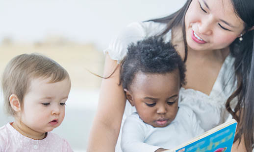 A woman reads to two very young children. One is sitting on her lap.