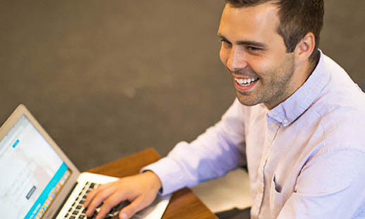 A man sits in a grey chair at a brown desk and types. He is smiling broadly and looking off to the left. He is wearing a light pink button-up shirt.