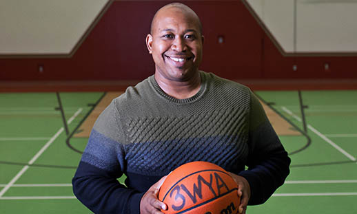 A man holding a basketball and wearing a sweater smiles at the camera. The ball is signed. He is in a basketball court with green flooring.