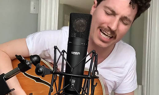 A man wearing a white t-shirt sits at a microphone and holds a brown guitar. His eyes are closed, and his mouth is open while he sings. 