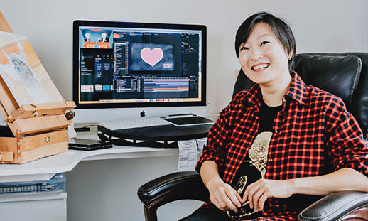 A woman wearing a plaid shirt sits at a computer. She is looking at the camera and her back is to her screen. She is holding a pen in her right hand.