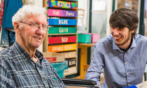 Two men sit at a table. One man is smiling at the other, who is in a wheelchair. They are in a classroom environment.