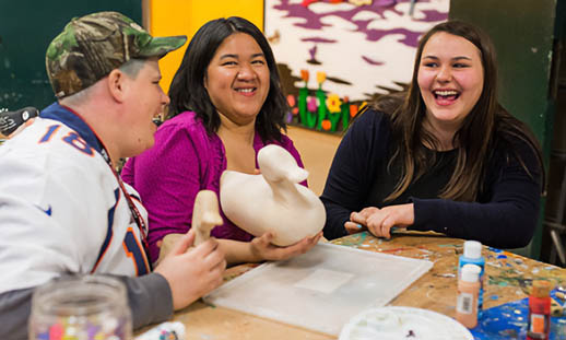 Two women and a man sit around a table. They are laughing and there are crafting supplies in front of them.