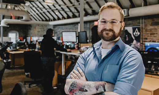 A man sits and leans into the camera. He is wearing a buttoned up shirt and white t-shirt. In the background, people can be seen working and there is a large Ubisoft sign. 