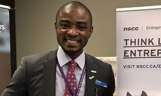 A man smiles broadly and looks at the camera. He is in a business suit and standing in front of two pull-up banners. One banner says Enactus and the other says NSCC Entrepreneurship.
