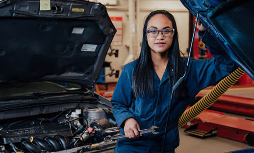 A young woman wearing blue coveralls stands in front of two cars with open engine bays. She is smiling and holds a wrench. She is in an automotive shop.