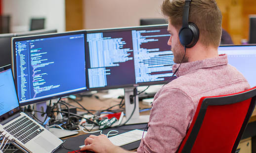 A man sits at a desk and looks at multiple computer screens. He is wearing headphones.