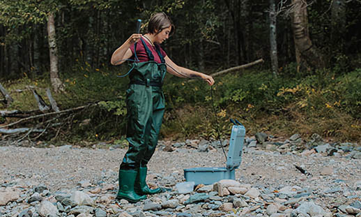 A woman in hip waders stands on a shoreline and takes a piece of equipment from a suitcase-style container.