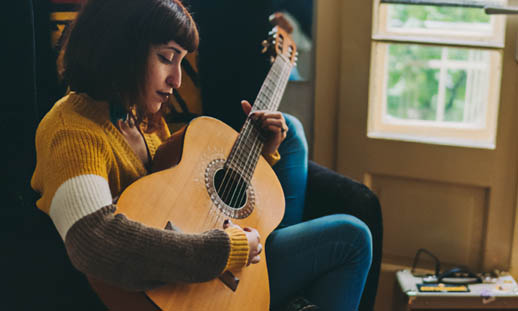 A woman in a yellow shirt strums a brown, acoustic guitar. There are two, open French doors seen in the background.