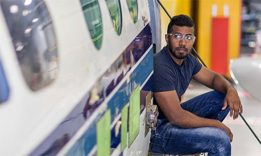 A man looks up while working underneath a plane. He is wearing blue work clothes and safety glasses. He is in a large, open workspace.