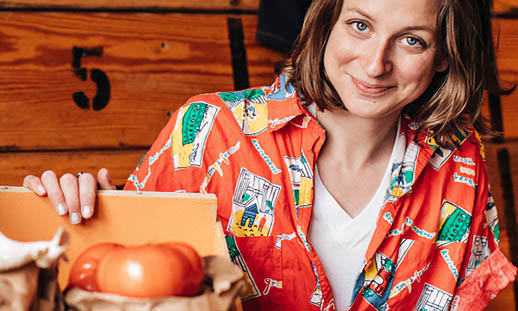 A woman in a patterned, orange shirt smiles at the camera. He hand is on a wooden crate containing produce.