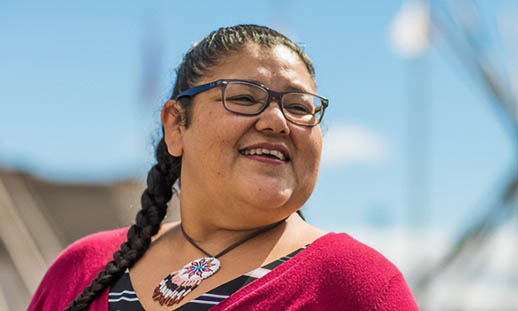 A woman in glasses smiles while looking off to the right of the image. She is wearing a maroon, button up sweater, a black and white striped t-shirt and a beaded necklace. He hair is in a braid. In the background, two teepee frames can be seen. 