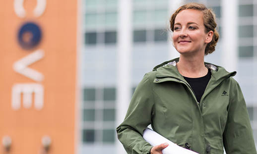 A woman in a green, zip-up jacket holds a rolled up map under her arm and walks in front of a building with the letters C O V E on it. 