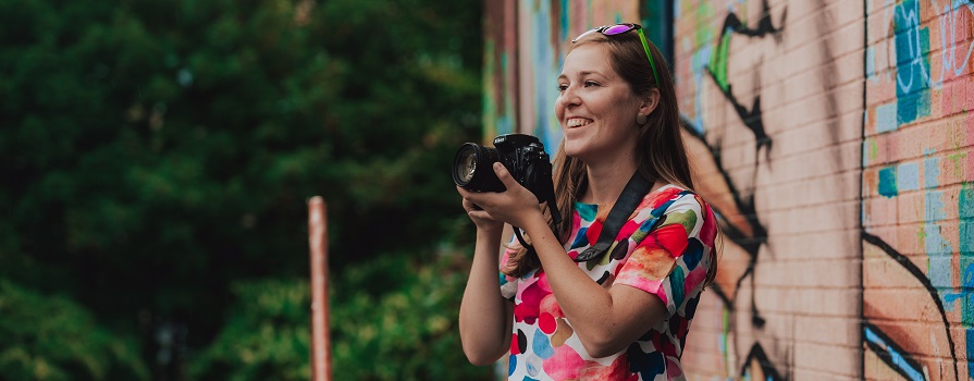 An NSCC photography student stands outdoors and smiles while holding a camera.