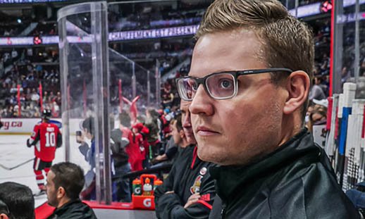 A man in glasses looks at something unseen to the left of the image. He is standing in the bench area of a large hockey stadium. People can be seen seated in front of him and playing hockey on the ice. There are many spectators in the background.