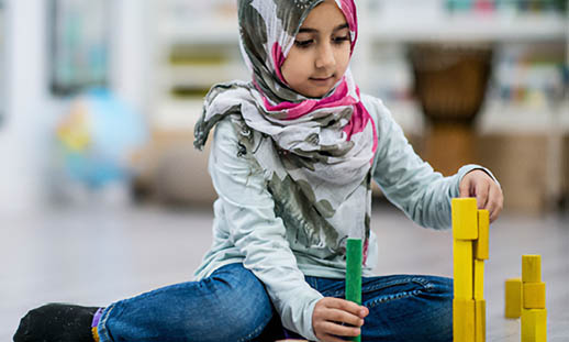 A young girl wearing a hijab sits on the floor playing with blocks.