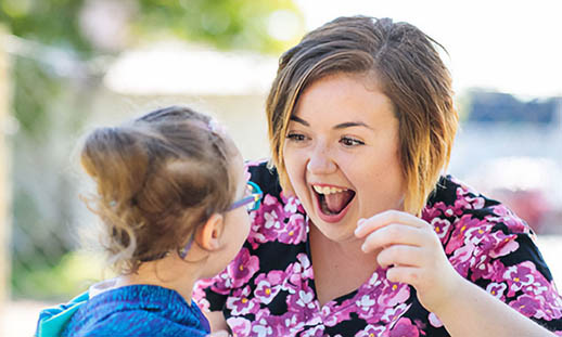 A young woman in a flowery, pink shirt smiles with an open mouth at a young child. They are seated at a picnic table in a park on a sunny day. 