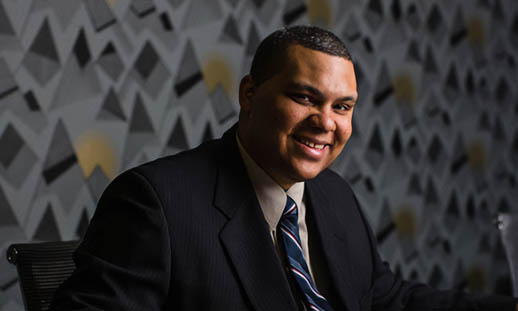 A man sits at a table and smiles at the camera. There is a laptop on the table. 