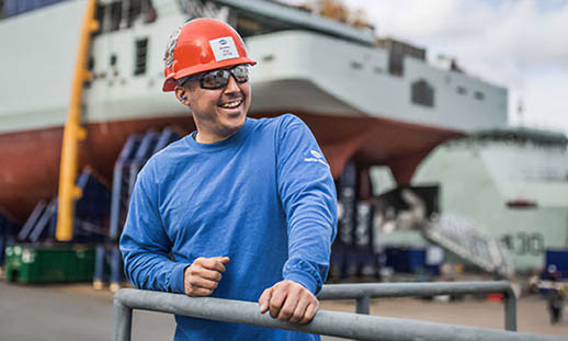 A man in a blue shirt, safety glasses and orange hardhat looks off to the top, right of the image. In the background there is a partially-constructed navy ship and a tall crane.