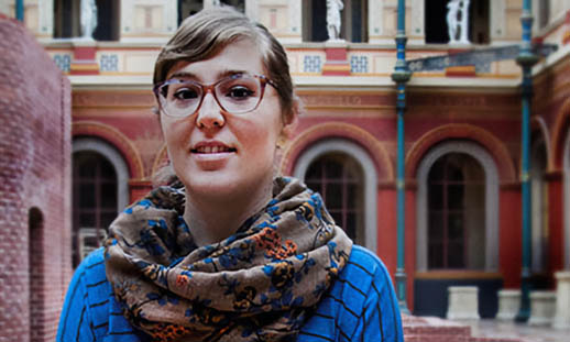 A woman wearing glasses, a blue sweater and scarf, stands in the atrium of a classically styled building with a glass domed roof.