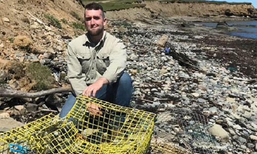A man crouches on a rocky shore. In front of him is a broken lobster trap.