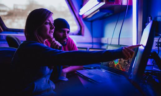 Two students sit near one another at a desk while looking at a monitor together. One student points at the monitor's screen.
