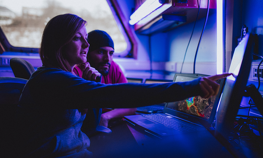 Two students sit near one another at a desk while looking at a monitor together. One student points at the monitor's screen.