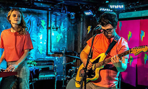 Two band members wearing orange shirts. The man is playing guitar and the woman is playing keyboards.