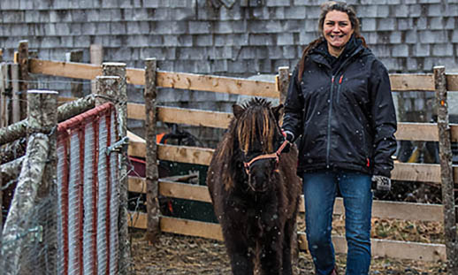 A woman in a black coat and jeans, walks a miniature horse between two wooden fences.