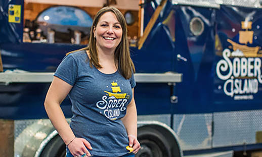 A woman in a blue t-shirt leans out the window of a blue food-truck-like structure and hands a customer a can of beer. The structure reads Sober Island Brewing.