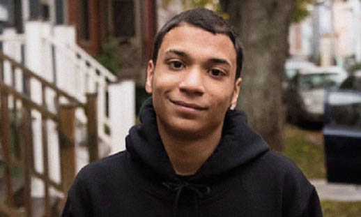 A head and shoulders shot of Unique Jones-MacKenzie. Unique is wearing a black hoodie and smiling slightly. A residential street can be seen behind him.
