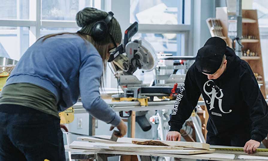 Two students work with wood in a shop-like classroom environment.