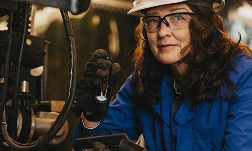 Christine MacKenzie inspects the weld on a piece of equipment with a flashlight. She is wearing a white hardhat, a blue smock, safety glasses and black gloves.