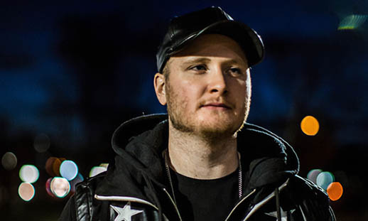 A young man wearing white pants, a black and white coat, black t-shirt and black hat, stands on a sidewalk and looks directly at the camera. It is night and the streetlights are illuminated. 