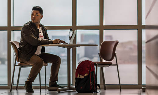 A man sits at a table and works on a laptop in front of a wall of windows.