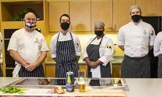 Six chefs wearing uniforms and facemasks stand behind a long counter. There are cupboards behind them. On the counter is a can of beer, a glass of beer and some produce.