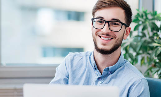 A man in glasses and a button up shirt smiles. He is in front of large windows and a plant. He appears to be working on a laptop.