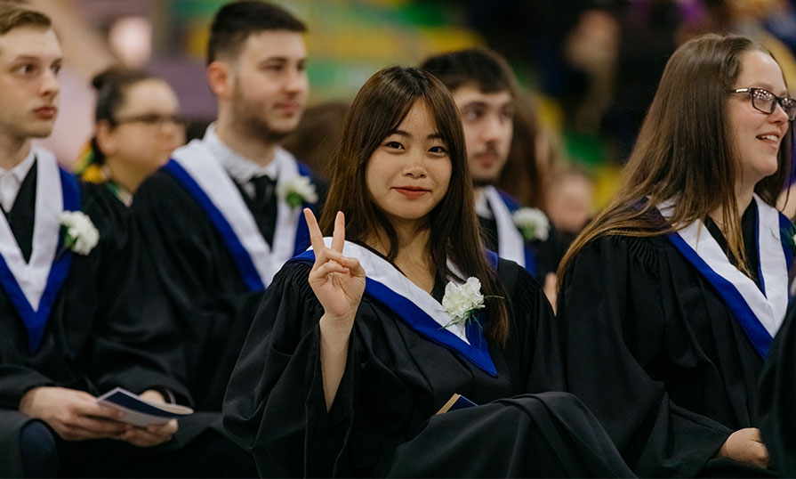 A young woman in a graduation gown smiles and makes a piece sign with her hands. She is seater among other graduates.