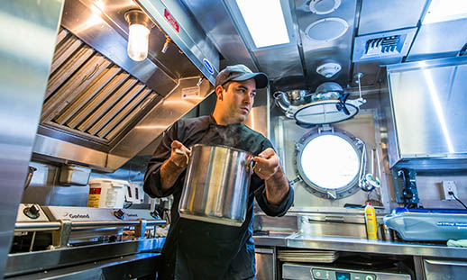 A man wearing a black chef's jacket and black ball cap, transfers a large, stainless steel pot across a kitchen. The kitchen is entirely stainless steel and small. A porthole window is open in the background.