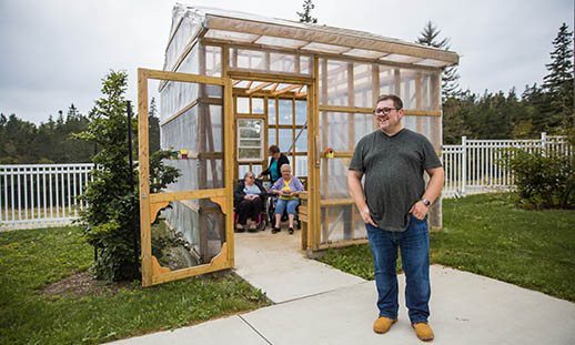 A man wearing a t-shirt, jeans and glasses, stands on the sidewalk outside of a greenhouse. In the greenhouse, several seated seniors can be seen.