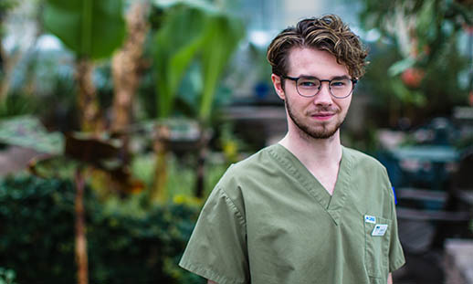 A young man wearing green scrubs and glasses looks into the camera. In the background lush, green plants can be seen.