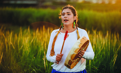 A woman wears a white shirt and holds a pepkwejete’maqn (Mi'kmaq drum). She is singing and beating the drum with a stick with a red end. Her hair is braided. There is a green field behind her.