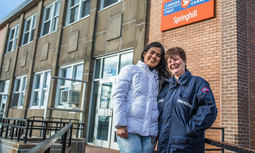 Two women stand in front of a Canada Post Office. One is wearing a white, winter jacket. The other woman is older and wears a Canada Post jacket. 