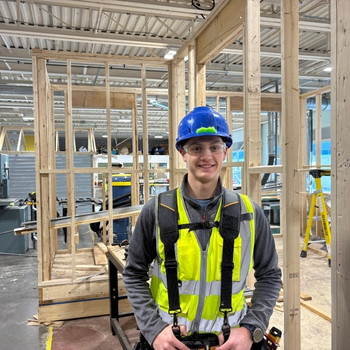 Ethan smiles in front of a wooden frame structure, wearing a hardhat and safety gear.