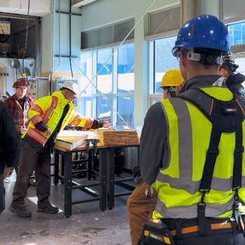 Instructor Andrew gives a carpentry demonstration in the workshop to a group of students, all wearing hardhats and reflective vests.