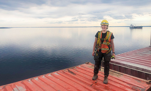 Olivia stands atop a red shipping container on the deck of a ship.